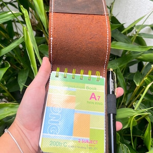 Puede incluir: Una cubierta de cuaderno de cuero marr&oacute;n con un borde cosido. La cubierta est&aacute; abierta para revelar un peque&ntilde;o cuaderno en espiral con un dise&ntilde;o colorido. El cuaderno est&aacute; etiquetado como "Composition Book A7 Twin Ring 2 Subject 6 mm x 14 d&ograve;ng MS: 301 200tr C". Un bol&iacute;grafo negro est&aacute; metido en la cubierta.