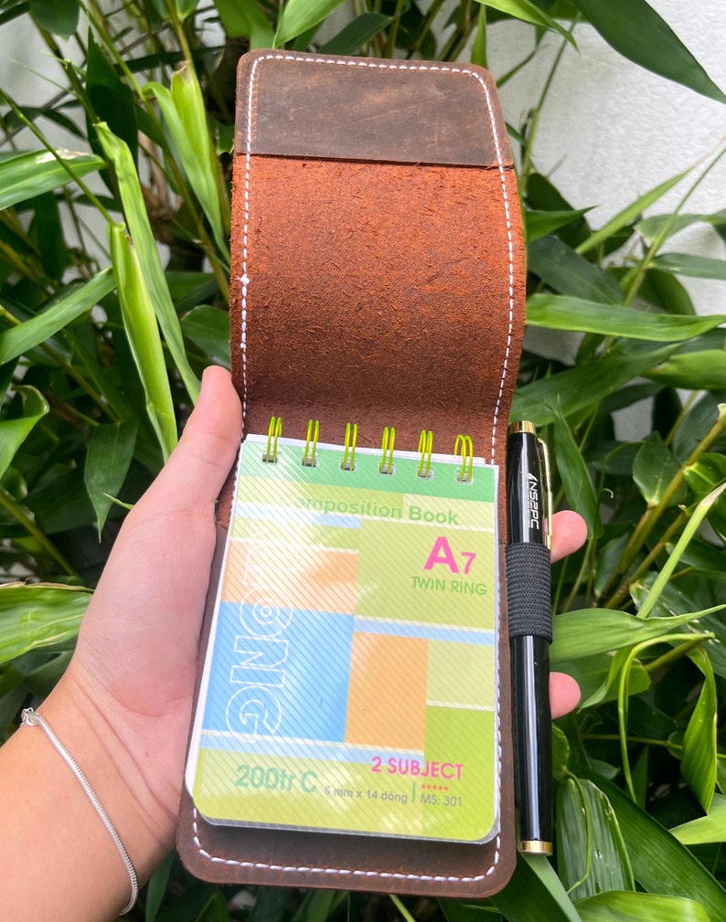 Puede incluir: Una cubierta de cuaderno de cuero marr&oacute;n con un borde cosido. La cubierta est&aacute; abierta para revelar un peque&ntilde;o cuaderno en espiral con un dise&ntilde;o colorido. El cuaderno est&aacute; etiquetado como "Composition Book A7 Twin Ring 2 Subject 6 mm x 14 d&ograve;ng MS: 301 200tr C". Un bol&iacute;grafo negro est&aacute; metido en la cubierta.