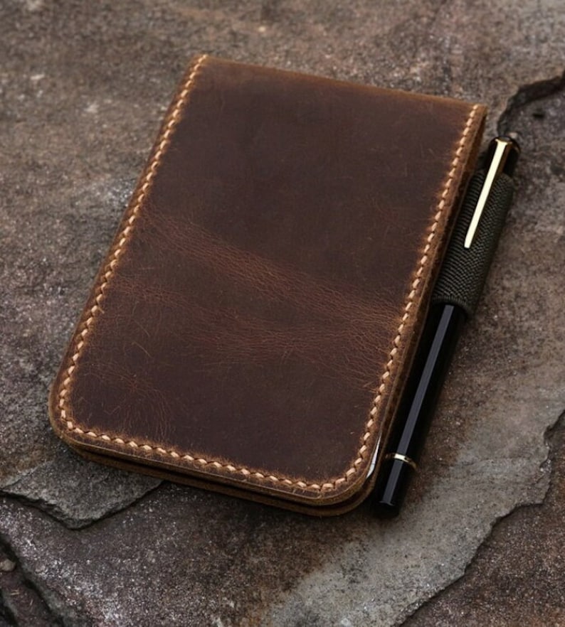 Puede incluir: Una cubierta de cuaderno de cuero marr&oacute;n con un portaplumas y un borde cosido.