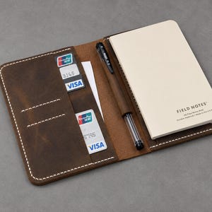May include: A brown leather notebook cover with white stitching, featuring two card slots containing Visa cards, a pen loop holding a black pen, and a cream-colored Field Notes memo book with the text Field Notes 48-Page Memo Book.