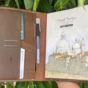 Może przedstawiać: Otwarty brązowy skórzany notatnik z długopisem, kartami kredytowymi i notatnikiem. Okładka notatnika zawiera tekst "Travel Stories" i akwarelową ilustrację europejskiego miasta z kopułami i budynkami.