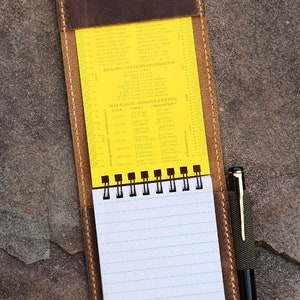 Puede incluir: Una cubierta de cuaderno de cuero marr&oacute;n con una regla amarilla y un cuaderno espiralado en el interior. La regla tiene medidas inglesas y m&eacute;tricas para escalas de mapas y medidas lineales. El cuaderno tiene papel rayado y el texto "Ret in the Rain" est&aacute; escrito en la parte inferior de la p&aacute;gina.