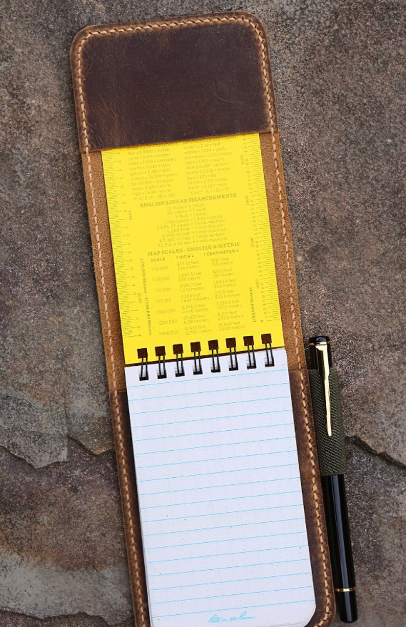 Puede incluir: Una cubierta de cuaderno de cuero marr&oacute;n con una regla amarilla y un cuaderno espiralado en el interior. La regla tiene medidas inglesas y m&eacute;tricas para escalas de mapas y medidas lineales. El cuaderno tiene papel rayado y el texto "Ret in the Rain" est&aacute; escrito en la parte inferior de la p&aacute;gina.