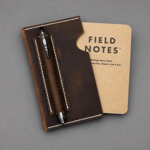 May include: A dark brown leather notebook cover with white stitching and a built-in pen loop holding a black mechanical pencil. A tan Field Notes 48-page memo book is inserted into the cover, showing the text Field Notes and Made in the U.S.A.