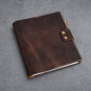 May include: A dark brown leather notebook cover featuring visible white stitching along the edges. The cover includes a secure flap closure with two brass-colored metal snap buttons and a small pen holder loop attached to the side of the spine.