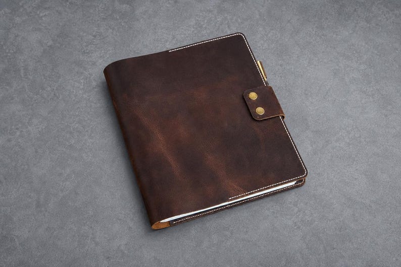 May include: A dark brown leather notebook cover featuring visible white stitching along the edges. The cover includes a secure flap closure with two brass-colored metal snap buttons and a small pen holder loop attached to the side of the spine.