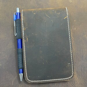 Puede incluir: Un cuaderno de cuero marr&oacute;n con costuras blancas y un bol&iacute;grafo azul.