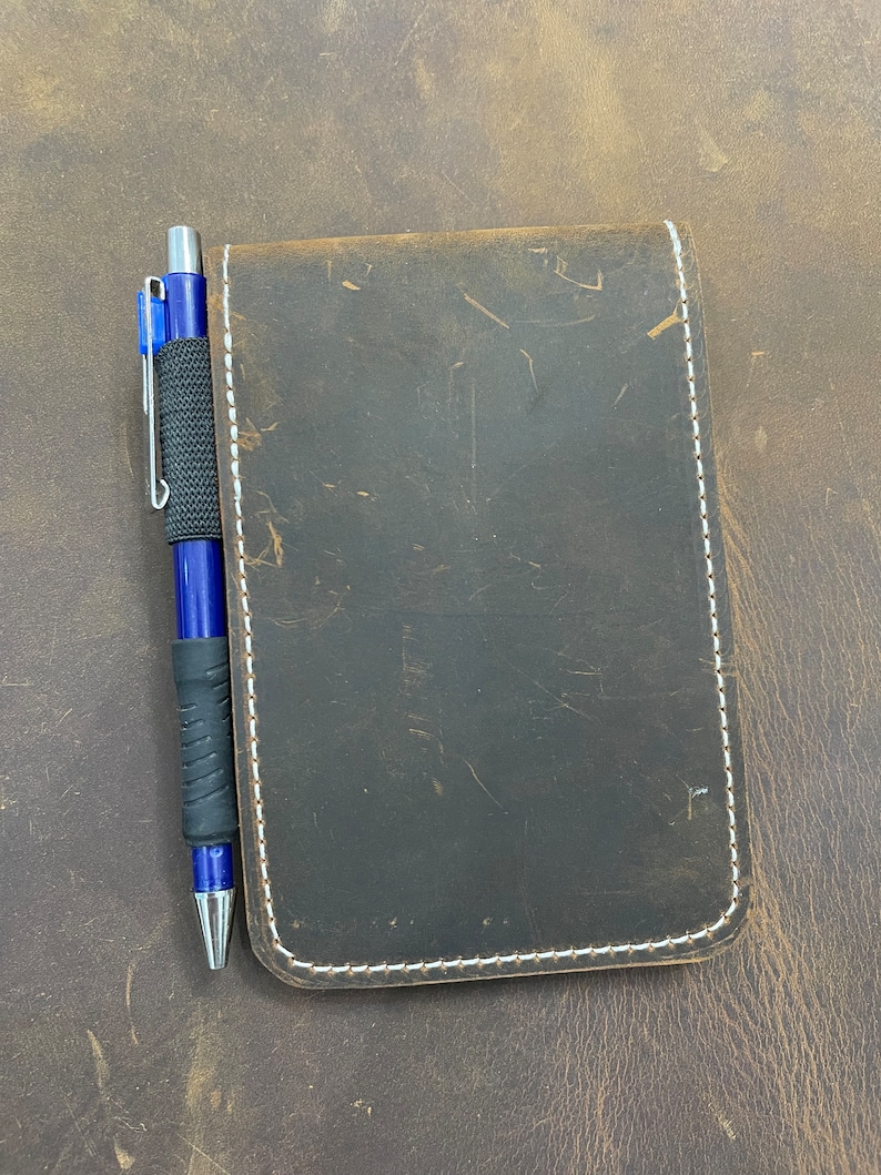 Puede incluir: Un cuaderno de cuero marr&oacute;n con costuras blancas y un bol&iacute;grafo azul.