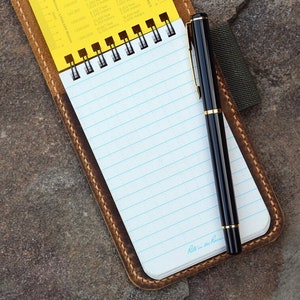Puede incluir: Una libreta de cuero marr&oacute;n con una regla amarilla y un bol&iacute;grafo negro. La libreta tiene una encuadernaci&oacute;n en espiral y p&aacute;ginas rayadas. El bol&iacute;grafo tiene un borde dorado.