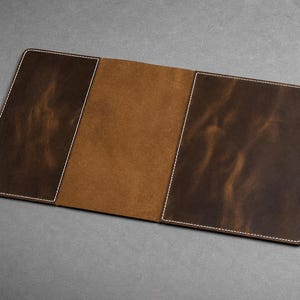 May include: A dark brown leather notebook cover featuring a smooth, matte finish with visible white stitching along the edges. The interior displays a lighter tan suede texture, and the exterior includes a side strap with two metal snap buttons.