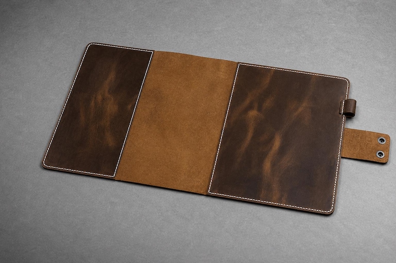 May include: A dark brown leather notebook cover featuring a smooth, matte finish with visible white stitching along the edges. The interior displays a lighter tan suede texture, and the exterior includes a side strap with two metal snap buttons.