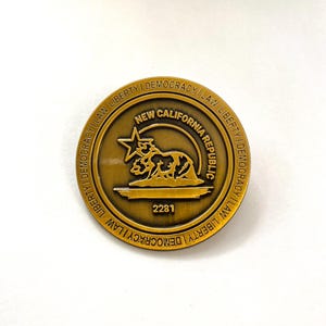 Könnte beinhalten: Eine goldfarbene Metallstifte mit den Worten "NEW CALIFORNIA REPUBLIC" und "LIBERTY | DEMOCRACY | LAW" am Rand. Die Mitte des Stifts zeigt einen Bären, der auf seinen Hinterbeinen steht, mit einem Stern darüber. Die Nummer "2281" ist unter dem Bären gedruckt.