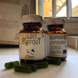 May include: Two brown glass bottles with white lids and labels. The label on the front bottle reads "Original Buffout 30 Tablets". The bottles are sitting on a white surface with several green capsules scattered around them.