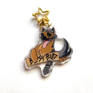 May include: A clear acrylic keychain featuring a cartoon German Shepherd dog wearing a hat with a brain design. The dog is holding a sign that says "Best Bud". The keychain has a gold star-shaped swivel clip.