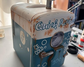 Puede incluir: Una réplica de máquina expendedora Quick Revive en azul y blanco con un acabado desgastado y oxidado. Presenta un letrero circular superior con un icono blanco sobre fondo rosa, y el texto "ICE COLD", "10¢" y "Quick Revive" en el frente.