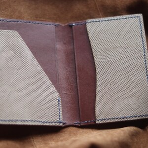 May include: Open leather wallet with a brown interior and light beige textured pockets. The edges are stitched with blue thread. The wallet is open, displaying the interior design.
