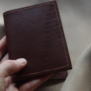 May include: A dark brown leather wallet with visible stitching and a textured surface. The wallet is rectangular and appears to be held by a person's hand. The background is a neutral beige color.