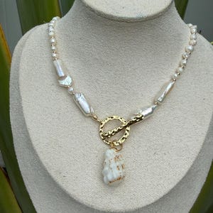 May include: A necklace featuring irregular white freshwater pearls of varying sizes, accented with small gold-toned beads. The piece includes a central gold-toned hammered toggle clasp with a single white textured shell pendant hanging below.