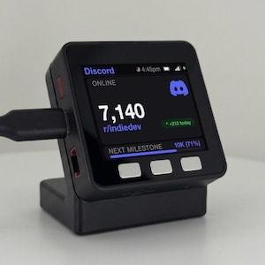 May include: A black square digital display device with three silver buttons at the bottom. The screen shows Discord, the text 7,140, r/indiedev, and a progress bar for the next milestone at 10K. A black charging cable is plugged into the side.