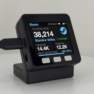 Op de afbeelding: Een zwart digitaal displayapparaat op een standaard, verbonden met een zwarte kabel. Het scherm toont Steam-gegevens voor Stardew Valley, waaronder 38.214 spelers, 14,4K beoordelingen, 12,2K positief en een voortgangsbalk naar de volgende mijlpaal op 50K (76%).