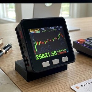 Może przedstawiać: Czarne, kwadratowe cyfrowe urządzenie typu ticker wyświetla wykres cen Bitcoina z tekstem 25821.58, Bitcoin oraz informacjami o dacie na ekranie. Urządzenie posiada trzy białe przyciski na dole i stoi na czarnej podstawce na drewnianym biurku.