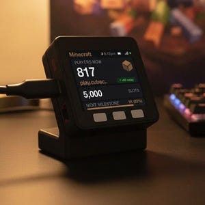 Puede incluir: Un dispositivo de pantalla digital negro sobre un soporte, conectado por un cable. La pantalla muestra estadísticas del servidor de Minecraft, incluyendo 817 jugadores, play.cubec..., 5.000 espacios y una barra de progreso al 1K (82%).