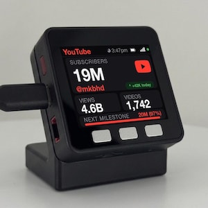 May include: A black digital subscriber counter device with a square screen displaying YouTube statistics including 19M subscribers for @mkbhd, 4.6B views, and 1,742 videos. The device features three silver buttons below the screen and a black charging cable.