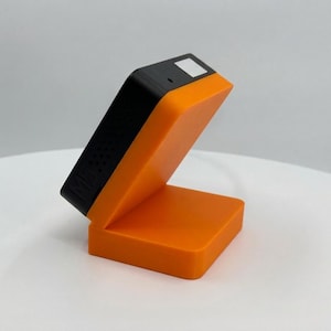 K&ouml;nnte beinhalten: Ein orangefarbenes und schwarzes elektronisches Ger&auml;t mit rechteckiger Basis und abgewinkelter Oberseite. Der obere Teil ist schwarz mit einem kleinen wei&szlig;en Quadrat und einem kleinen Loch. Das Ger&auml;t steht auf einer wei&szlig;en Oberfl&auml;che.