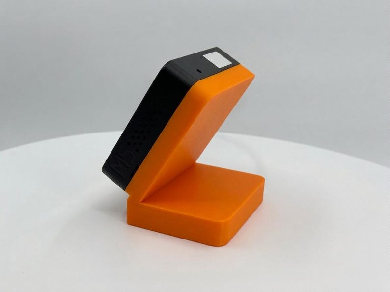 K&ouml;nnte beinhalten: Ein orangefarbenes und schwarzes elektronisches Ger&auml;t mit rechteckiger Basis und abgewinkelter Oberseite. Der obere Teil ist schwarz mit einem kleinen wei&szlig;en Quadrat und einem kleinen Loch. Das Ger&auml;t steht auf einer wei&szlig;en Oberfl&auml;che.