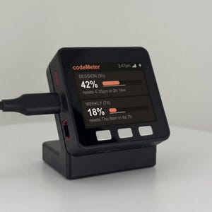 K&ouml;nnte beinhalten: Ein schwarzes codeMeter-Ger&auml;t mit einem Bildschirm, der Nutzungsstatistiken anzeigt. Der Bildschirm zeigt "SESSION (5h)" mit 42% und "WEEKLY (7d)" mit 18% an. Das Ger&auml;t ist an ein schwarzes Ladekabel angeschlossen und steht in einer schwarzen Basis.