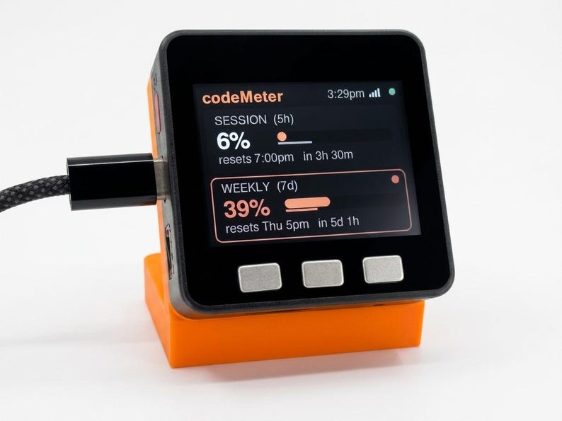 K&ouml;nnte beinhalten: Ein schwarzes und orangefarbenes digitales Ger&auml;t mit der Aufschrift "codeMeter" auf dem Bildschirm. Der Bildschirm zeigt Fortschrittsbalken f&uuml;r Sitzungen und Wochen an. Das Ger&auml;t ist mit einem schwarzen Kabel angeschlossen und steht auf einem orangefarbenen St&auml;nder.