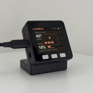 K&ouml;nnte beinhalten: Ein schwarzes CodeMeter-Ger&auml;t mit einem Bildschirm, der Daten anzeigt und mit einem schwarzen Kabel verbunden ist. Der Bildschirm zeigt den Text "codeMeter", "SESSION (5h)", "42%" und "WEEK 18%" sowie weitere Details. Das Ger&auml;t steht auf einer wei&szlig;en Oberfl&auml;che.