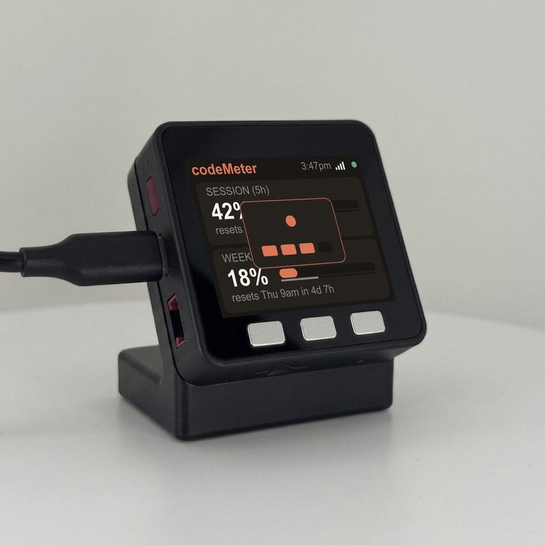 K&ouml;nnte beinhalten: Ein schwarzes CodeMeter-Ger&auml;t mit einem Bildschirm, der Daten anzeigt und mit einem schwarzen Kabel verbunden ist. Der Bildschirm zeigt den Text "codeMeter", "SESSION (5h)", "42%" und "WEEK 18%" sowie weitere Details. Das Ger&auml;t steht auf einer wei&szlig;en Oberfl&auml;che.