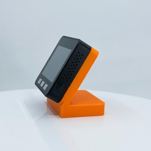 K&ouml;nnte beinhalten: Ein schwarzes elektronisches Ger&auml;t mit Bildschirm, das auf einem orangefarbenen St&auml;nder steht. Das Ger&auml;t hat drei Tasten und kleine L&ouml;cher an der Seite. Der St&auml;nder ist leuchtend orange und abgewinkelt, um das Ger&auml;t aufrecht zu halten.