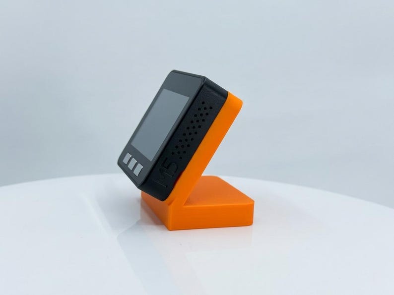 K&ouml;nnte beinhalten: Ein schwarzes elektronisches Ger&auml;t mit Bildschirm, das auf einem orangefarbenen St&auml;nder steht. Das Ger&auml;t hat drei Tasten und kleine L&ouml;cher an der Seite. Der St&auml;nder ist leuchtend orange und abgewinkelt, um das Ger&auml;t aufrecht zu halten.