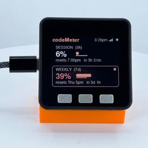 K&ouml;nnte beinhalten: Ein schwarzes digitales Ger&auml;t mit der Aufschrift "codeMeter" auf dem Bildschirm, das Sitzungs- und Wocheninformationen anzeigt. Das Ger&auml;t steht auf einem orangefarbenen St&auml;nder und ist mit einem Kabel verbunden.