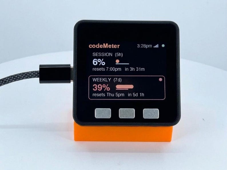 K&ouml;nnte beinhalten: Ein schwarzes digitales Ger&auml;t mit der Aufschrift "codeMeter" auf dem Bildschirm, das Sitzungs- und Wocheninformationen anzeigt. Das Ger&auml;t steht auf einem orangefarbenen St&auml;nder und ist mit einem Kabel verbunden.