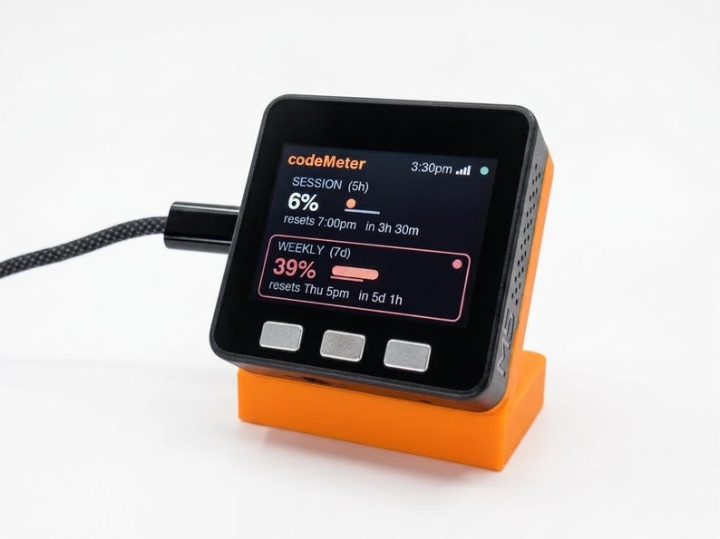 K&ouml;nnte beinhalten: Ein schwarzes und orangefarbenes digitales Ger&auml;t mit einem Bildschirm, der Daten anzeigt, darunter der Text "codeMeter", "SESSION" und "WEEKLY". Das Ger&auml;t ist angeschlossen und ruht auf einem orangefarbenen St&auml;nder. Der Bildschirm zeigt Prozents&auml;tze und die verbleibende Zeit an.