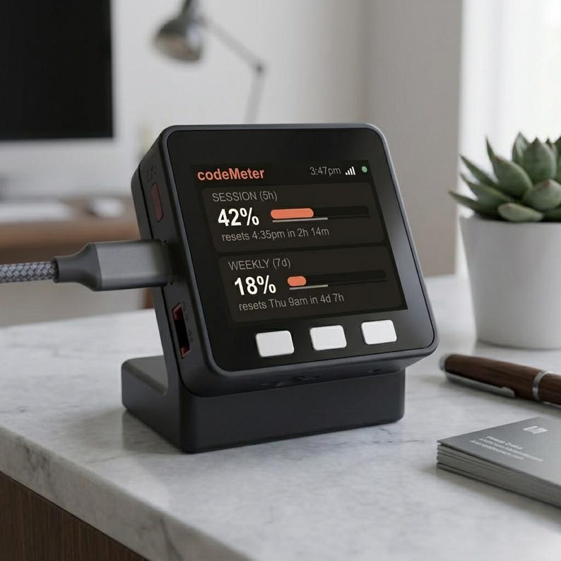 K&ouml;nnte beinhalten: Ein schwarzes digitales Ger&auml;t mit einem Bildschirm, der Daten anzeigt, verbunden mit einem Ladekabel. Der Bildschirm zeigt den Text "codeMeter" und Datenbalken mit Prozenten und Restzeit an. Das Ger&auml;t steht auf einer schwarzen Basis.