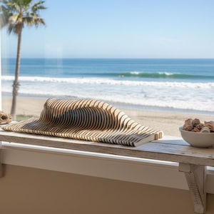 May include: A wooden wave sculpture with horizontal lines sits on a white shelf. A small bowl of seashells and a piece of driftwood are also on the shelf. The ocean and a palm tree are visible through the window.