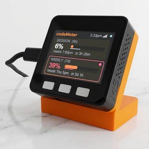 May include: A black and orange electronic device with a screen displaying data, including the text "codeMeter". The device is on an orange stand. A black cable is connected to the device.