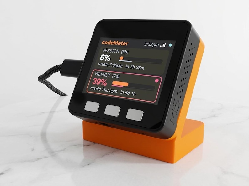 K&ouml;nnte beinhalten: Ein schwarzes und orangefarbenes elektronisches Ger&auml;t mit einem Bildschirm, der Daten anzeigt, einschlie&szlig;lich des Textes "codeMeter". Das Ger&auml;t steht auf einem orangefarbenen St&auml;nder. Ein schwarzes Kabel ist mit dem Ger&auml;t verbunden.