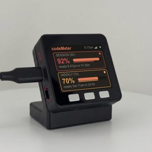 K&ouml;nnte beinhalten: Ein schwarzes digitales Ger&auml;t mit einem Bildschirm, der Nutzungsstatistiken anzeigt, einschlie&szlig;lich Prozents&auml;tzen und R&uuml;cksetzzeiten. Das Ger&auml;t ist mit einem Ladekabel verbunden und steht in einem passenden schwarzen St&auml;nder. Der Bildschirm zeigt "codeMeter".