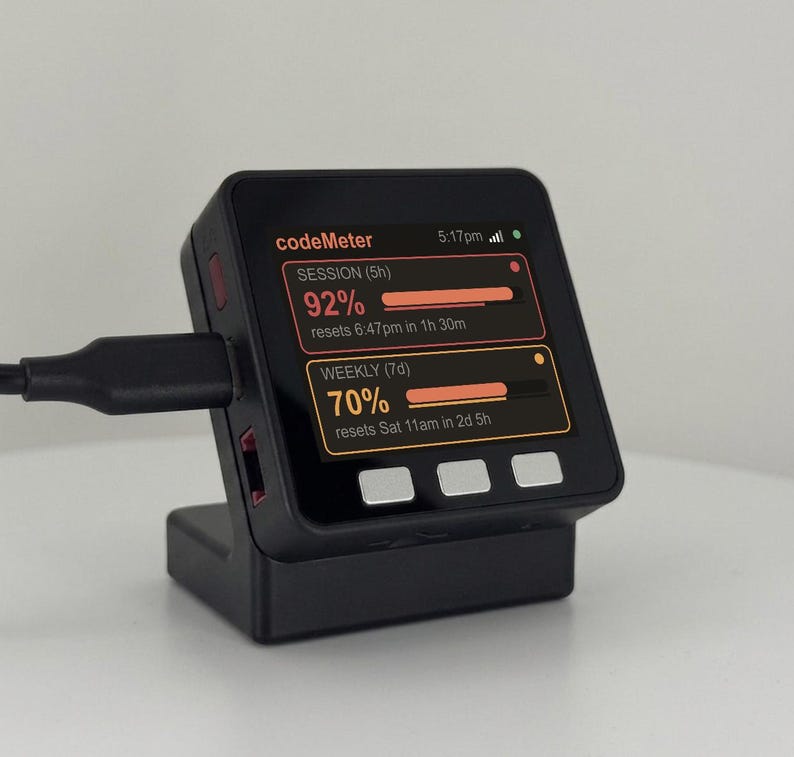K&ouml;nnte beinhalten: Ein schwarzes digitales Ger&auml;t mit einem Bildschirm, der Nutzungsstatistiken anzeigt, einschlie&szlig;lich Prozents&auml;tzen und R&uuml;cksetzzeiten. Das Ger&auml;t ist mit einem Ladekabel verbunden und steht in einem passenden schwarzen St&auml;nder. Der Bildschirm zeigt "codeMeter".