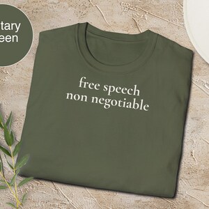 Puede incluir: Camiseta verde militar con las palabras "free speech non negotiable" en blanco. La camiseta está hecha de un material suave y está sobre una superficie texturizada. Un sombrero blanco y una rosa están en el fondo.