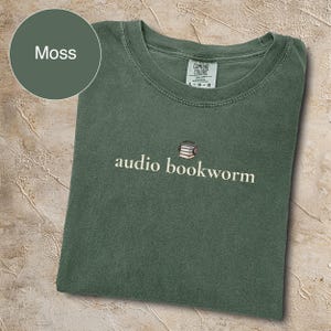 May include: A moss green t-shirt with the words "audio bookworm" in white script. The design includes a small graphic of a stack of books with headphones. The shirt is folded and has a Comfort Colors tag.