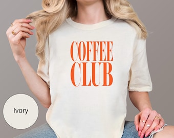May include: Ivory-colored t-shirt with the words "COFFEE CLUB" in bold orange letters. The shirt is a casual style, likely made of soft fabric. The text is centered on the front of the shirt.