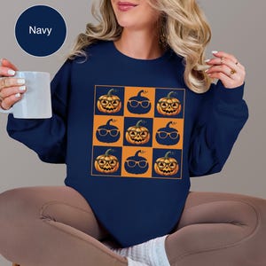 May include: Navy blue sweatshirt featuring a grid of nine orange pumpkins wearing glasses against a blue and orange checkered background. The pumpkins have smiling faces. A navy blue circle with the word "Navy" is in the upper left corner.