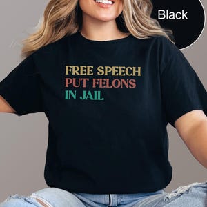 Puede incluir: Camiseta negra con el texto "FREE SPEECH PUT FELONS IN JAIL" en letras amarillas, rojas y verdes. La camiseta es de color negro liso y tiene cuello redondo.