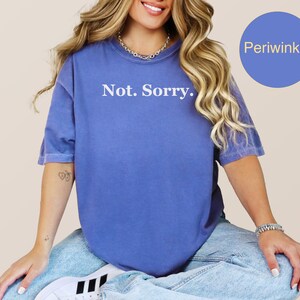 May include: A periwinkle blue t-shirt with the words "Not. Sorry." printed in white. The shirt is a solid color and has short sleeves. The model is wearing blue jeans and white sneakers.